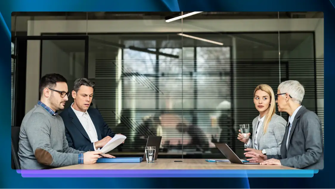 Four people at a conference room table. Two are looking at papers, two are on laptops and one is drinking water.