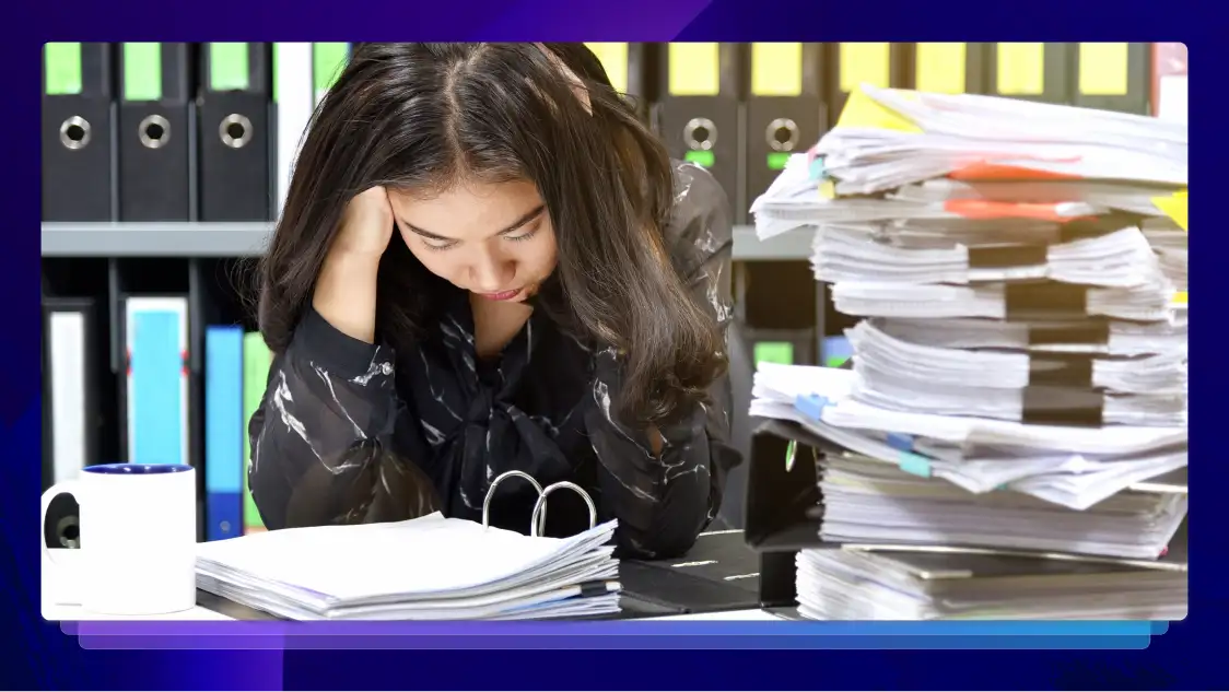 A woman sits with her head in her hand at a desk. She is surrounded by a coffee mug and stacks of papers.