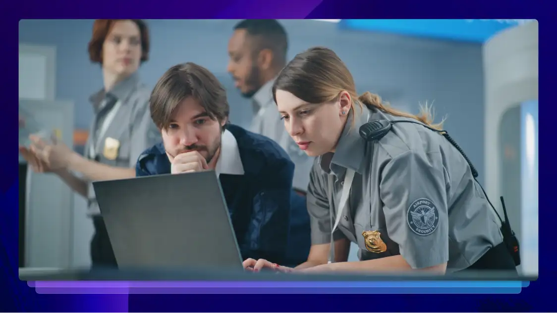A police officer in uniform and a law enforcement official in a blue coat work on a laptop with bystanders in the background.