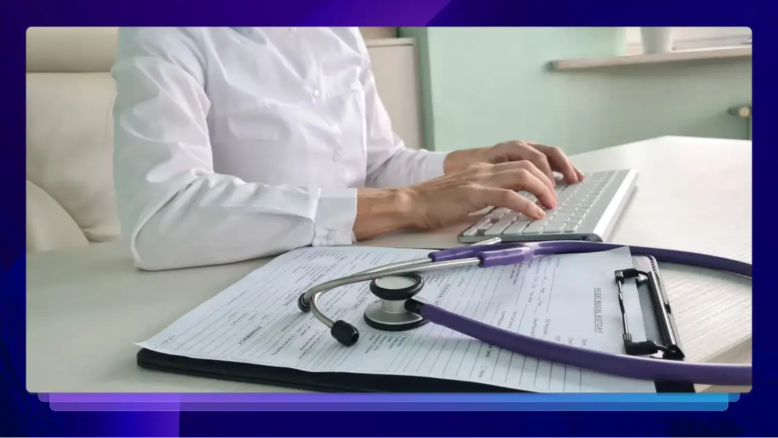 A medical professional typing on a keyboard at a desk with a stethoscope and medical documents placed on a clipboard in the foreground