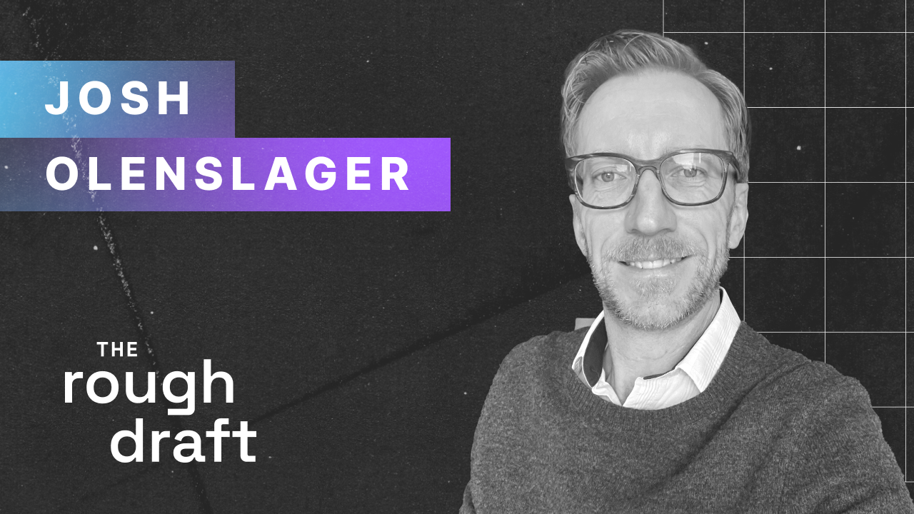 Professional headshot portrait of a man in blazer smiling against neutral gray background