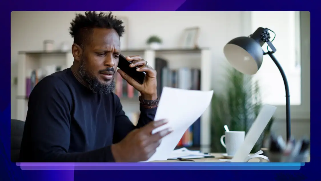 Man on phone reviewing documents, working from office with laptop, coffee cup, and desk lamp in the background.
