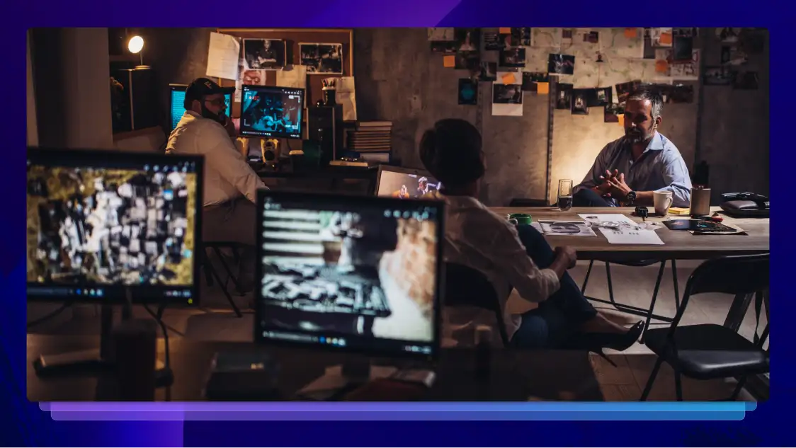 Three police officers sit at two different desks working on a case in a dark room surrounded by computers, photographs, and documents.
