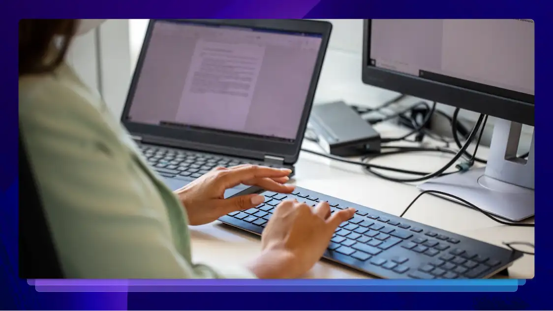 Person typing on a keyboard while working on a document with two computer screens on a desk.