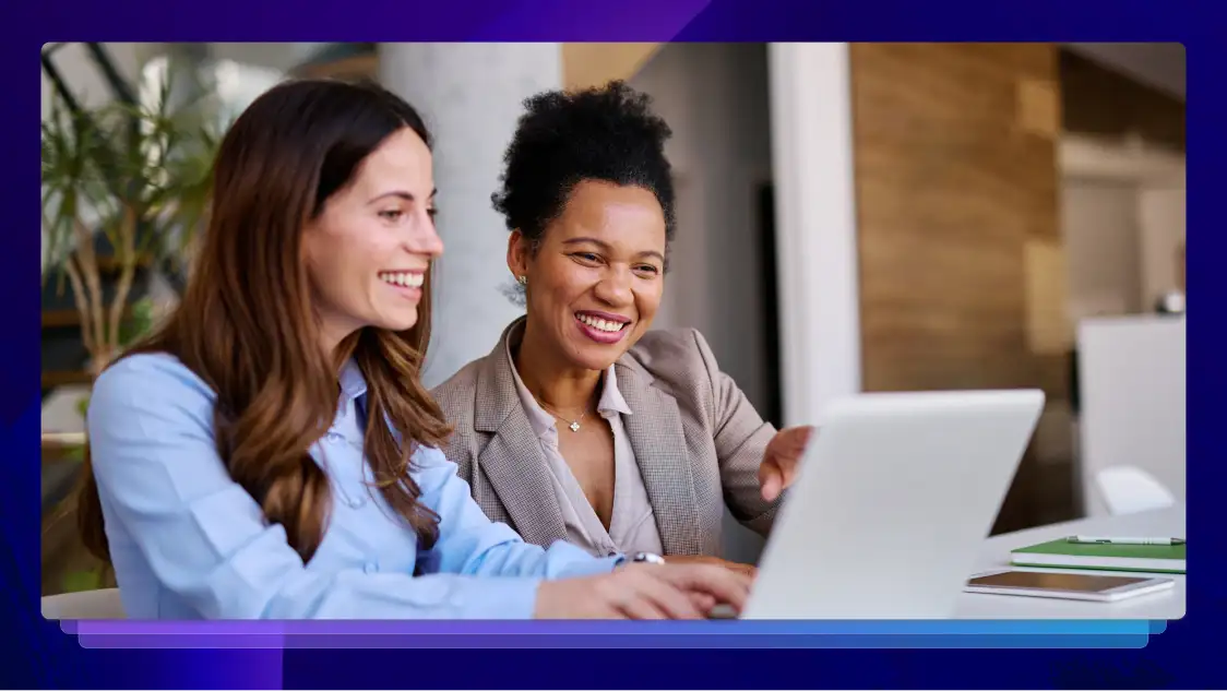 Two professional women smiling and working together on a laptop in a modern office setting.