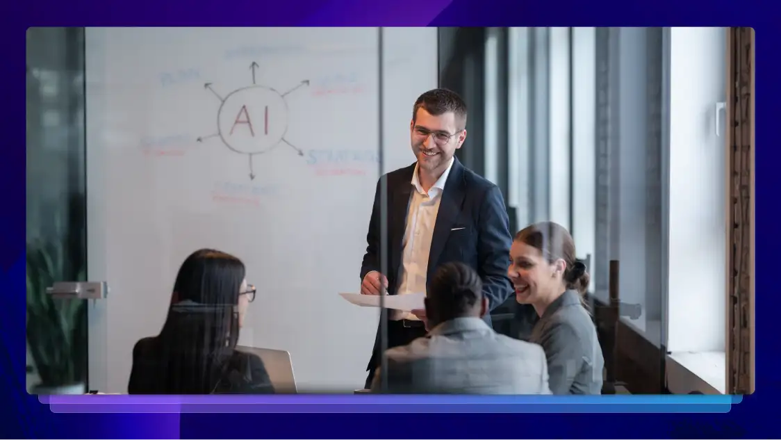 A group of people are in a meeting behind a glass wall. A man is standing and smiling, and there is a whiteboard with “AI” written on it behind him.