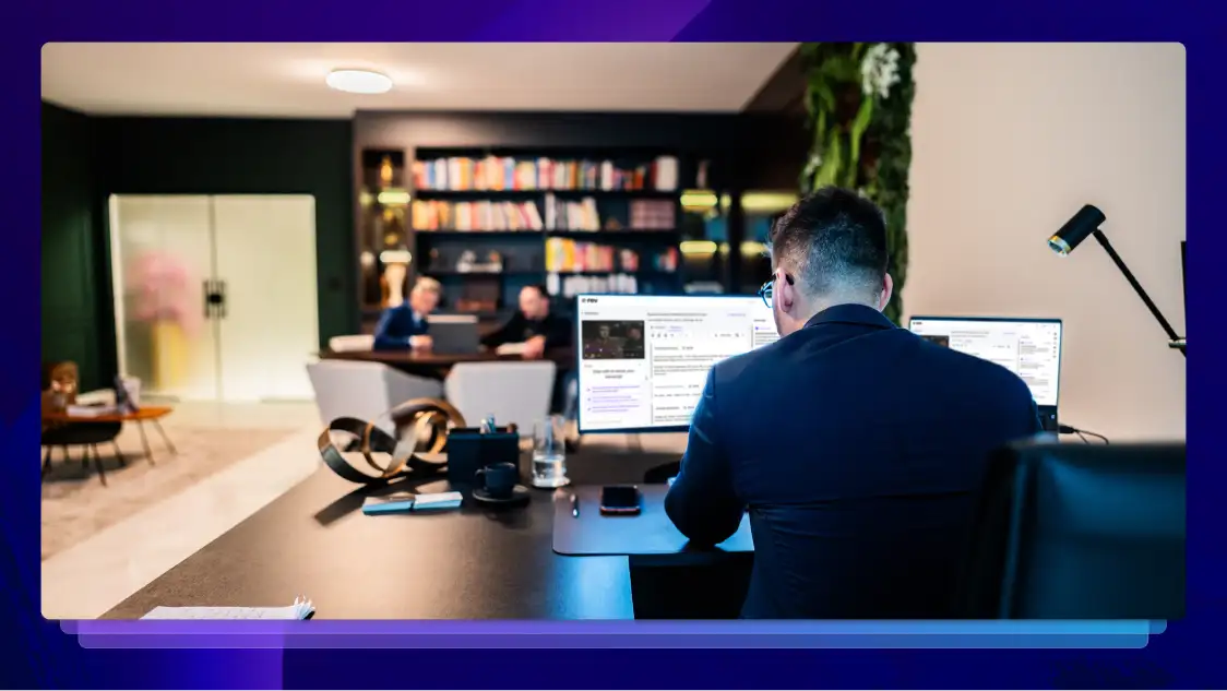 A man sits at a desk looking at the Rev AI and Transcription platform on two monitors. There are several other people sitting at another desk in the same room.