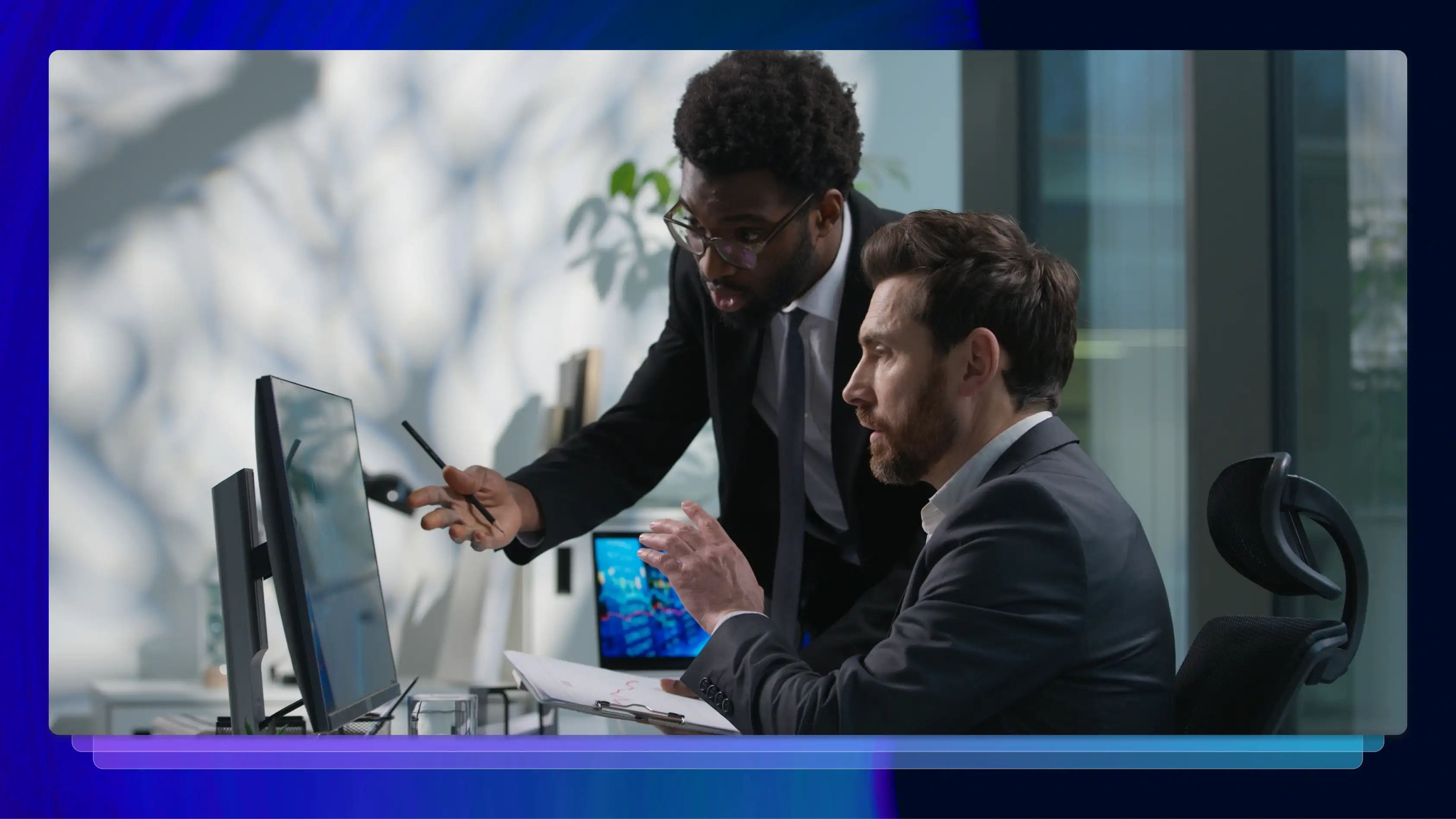 Two men and suits talk to each other and point at a computer screen in front of them.