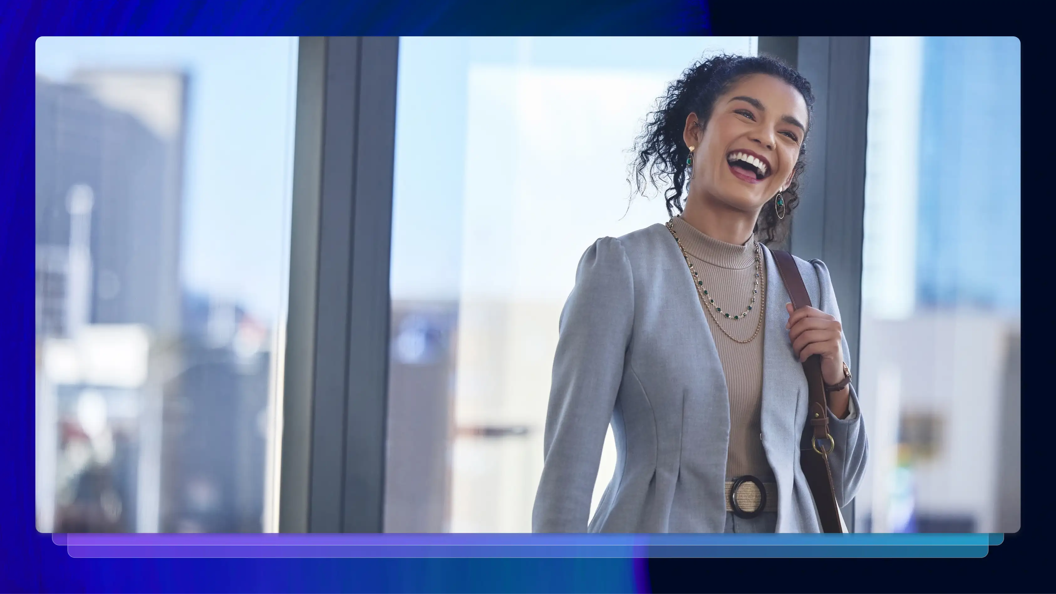 A woman in a blazer smiles with her mouth open. She is standing in front of several large windows, and buildings are visible.