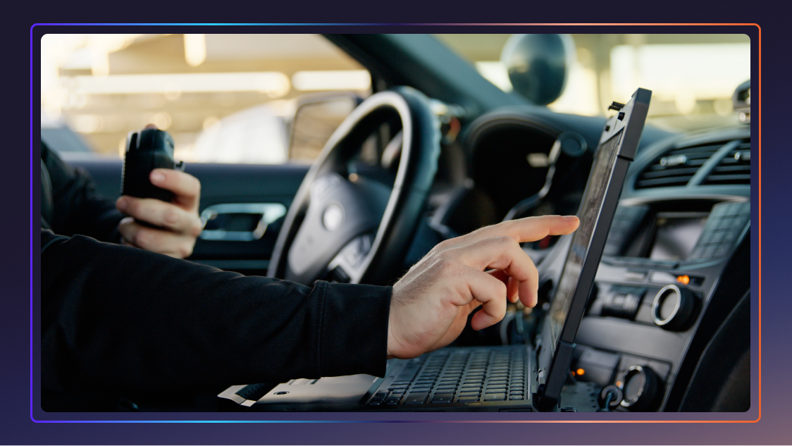 Close-up of a police officer's hands inside a police car. They are touching a laptop screen with one hand and holding a walkie-talkie with the other.