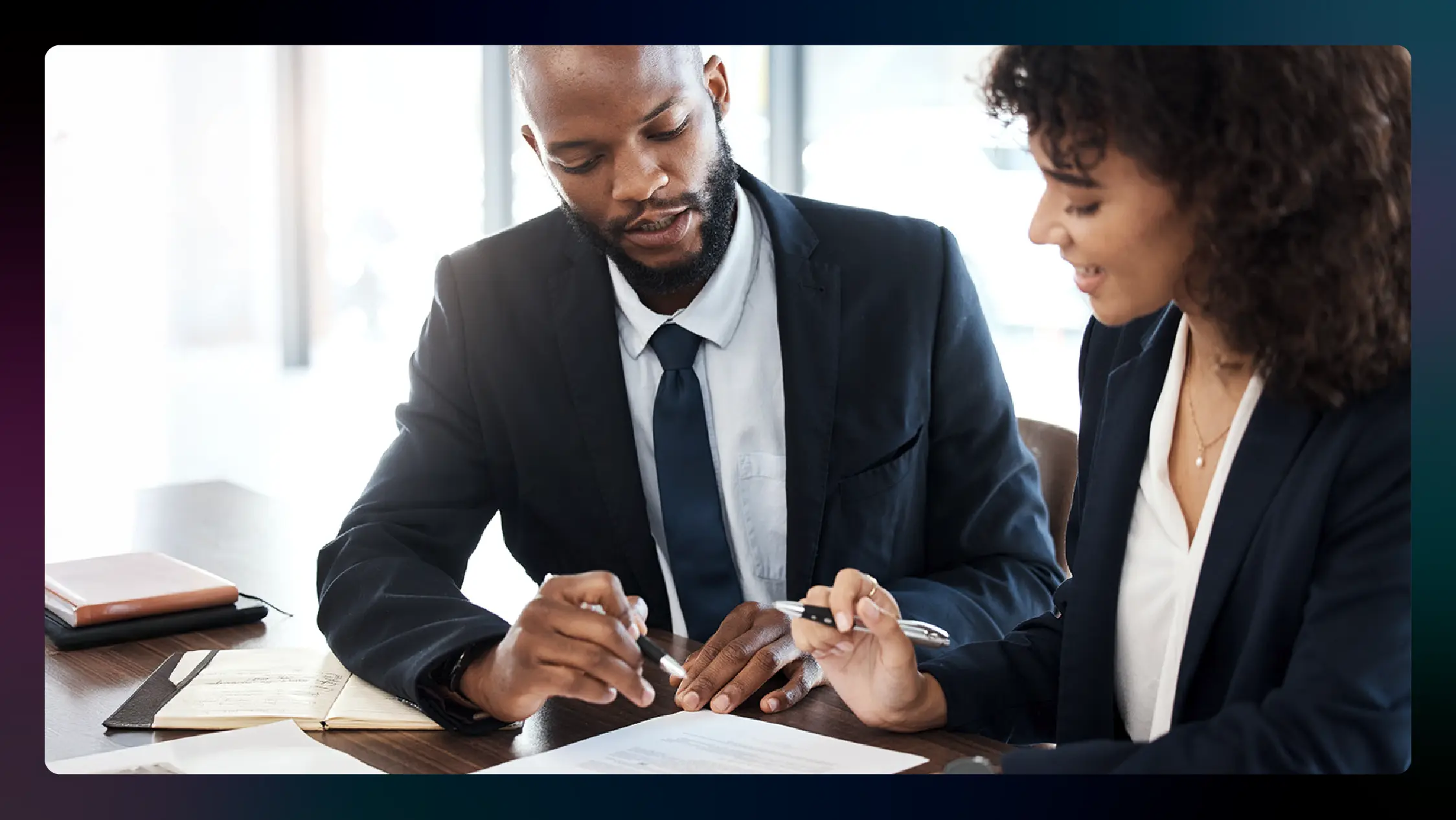 A man and woman in suits examine legal paperwork while seated at a conference table.