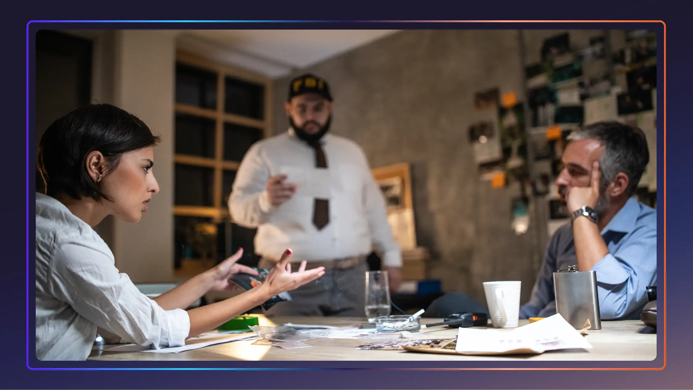 Two detectives sit across from each other and talk at a table full of evidence. One detective stands nearby and looks at a piece of paper.