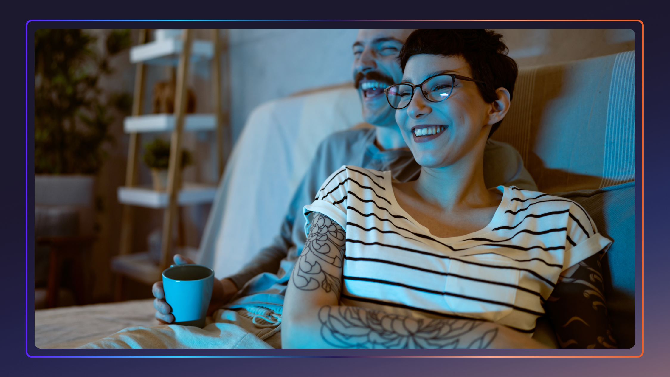 A man and a woman sit and laugh on a couch while watching a movie. The woman has tattoos on her arm.