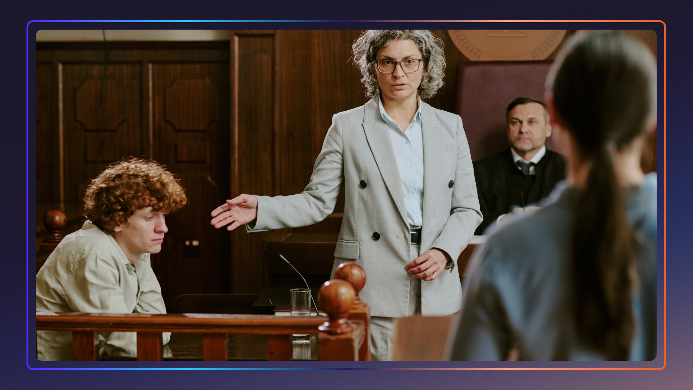 A lawyer stands and gestures to a witness on the stand. A judge stands behind her and a woman stands in front of her.