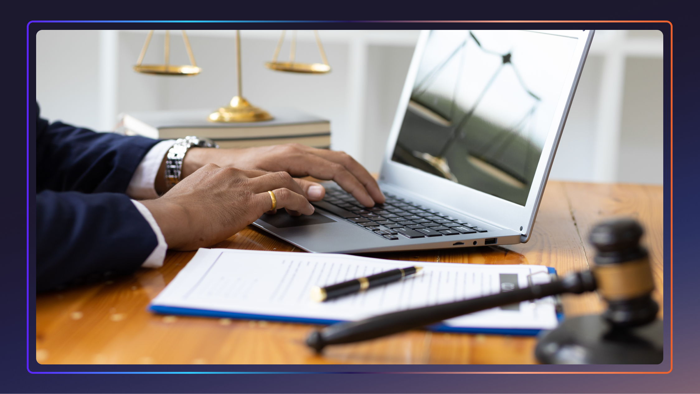 A man’s hands type on a laptop that is sitting on a desk. There is a gavel, a clipboard, and a Scale of Justice also on the desk.