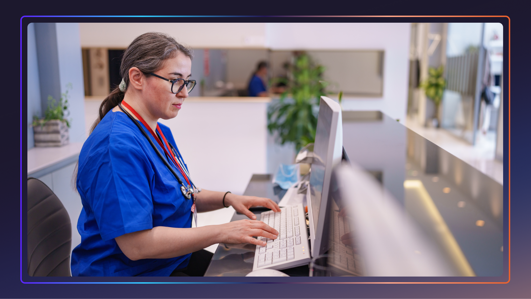 A woman in scrubs sits and types at a computer inside a hospital.