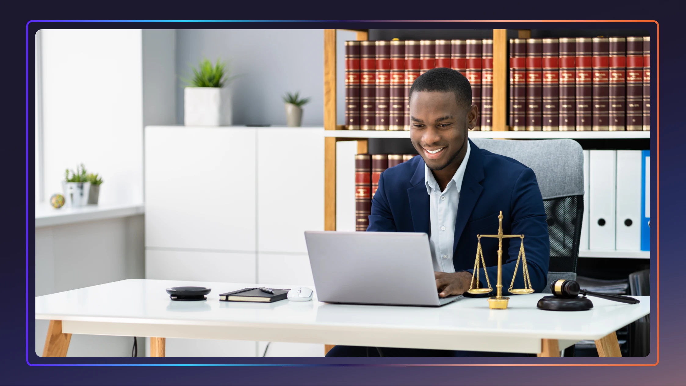 A young lawyer sits at his desk smiling and typing on his laptop.