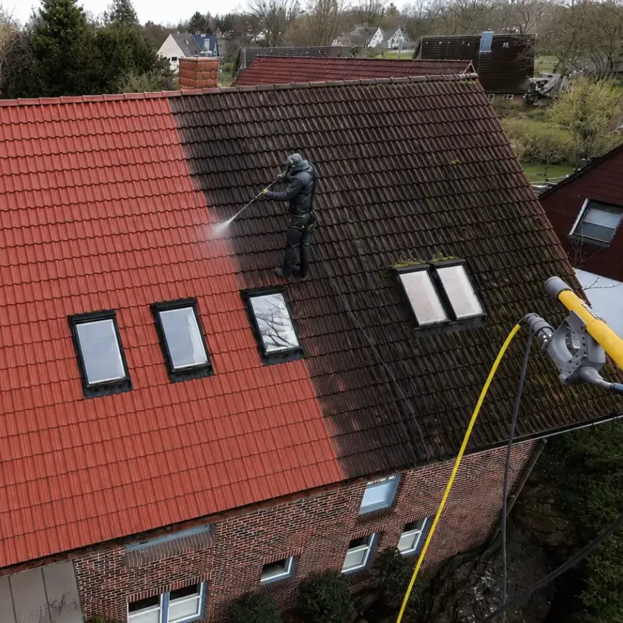 Person reinigt mit Hochdruckreiniger das Dach eines Hauses, halb sauber, halb schmutzig.