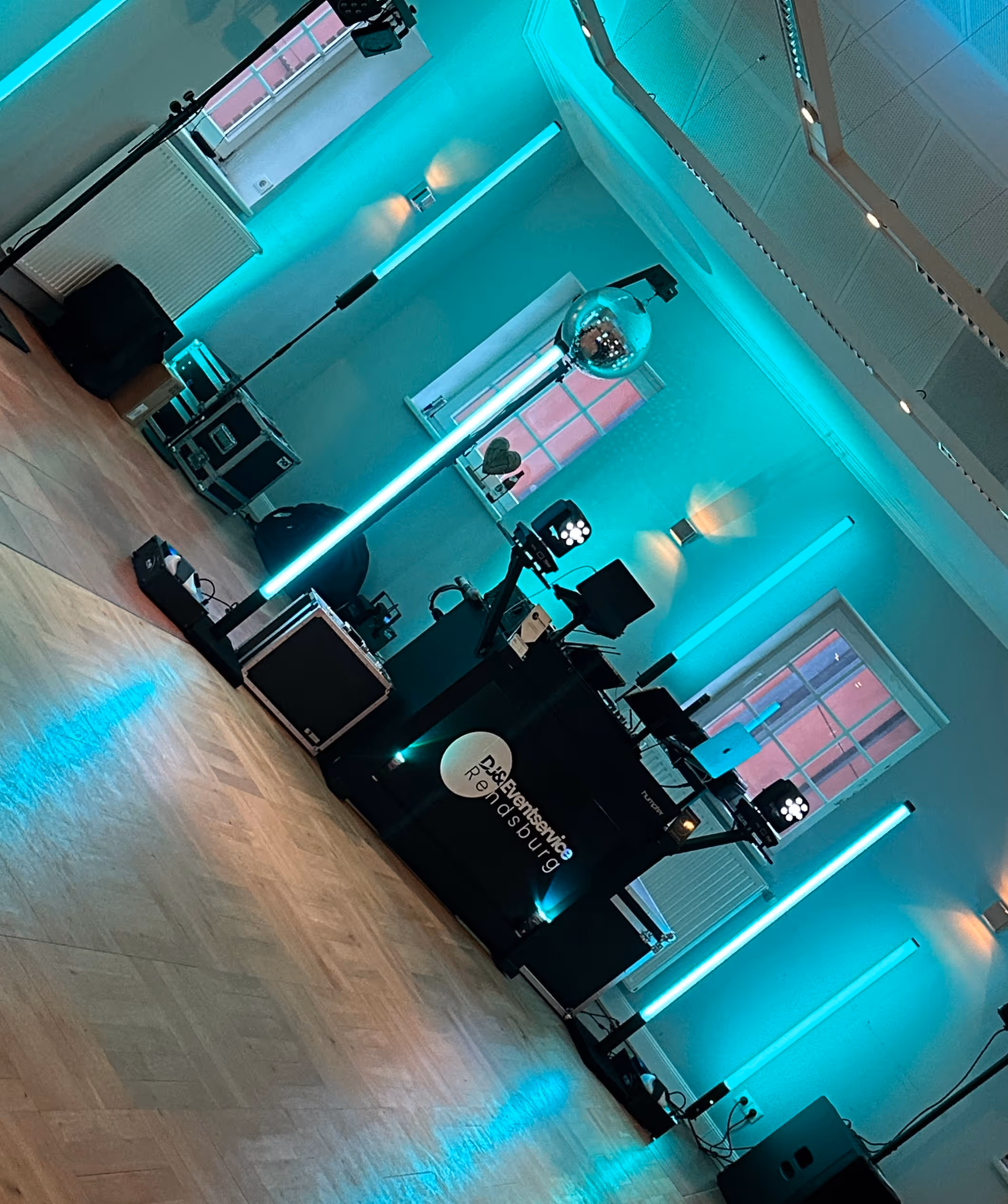 DJ-Setup in einem Raum mit Holzboden und türkisfarbener Beleuchtung, einschließlich Disco-Kugel und Lautsprechern.