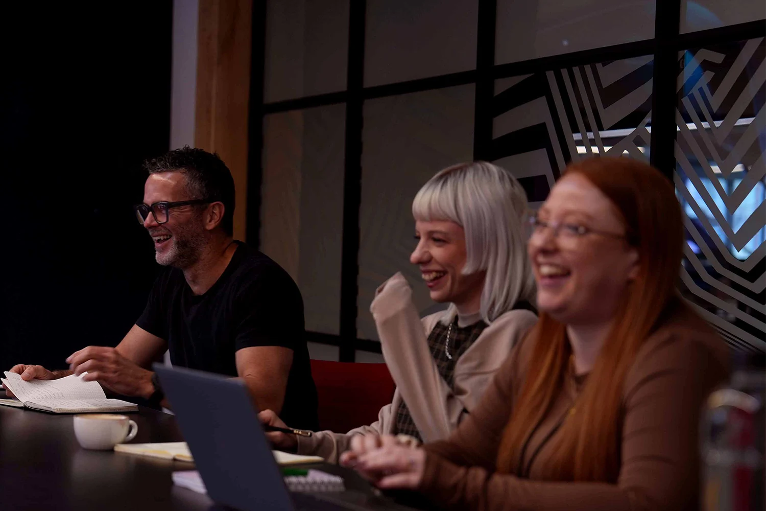 Three people sitting at a table in a modern office, laughing and enjoying a conversation.