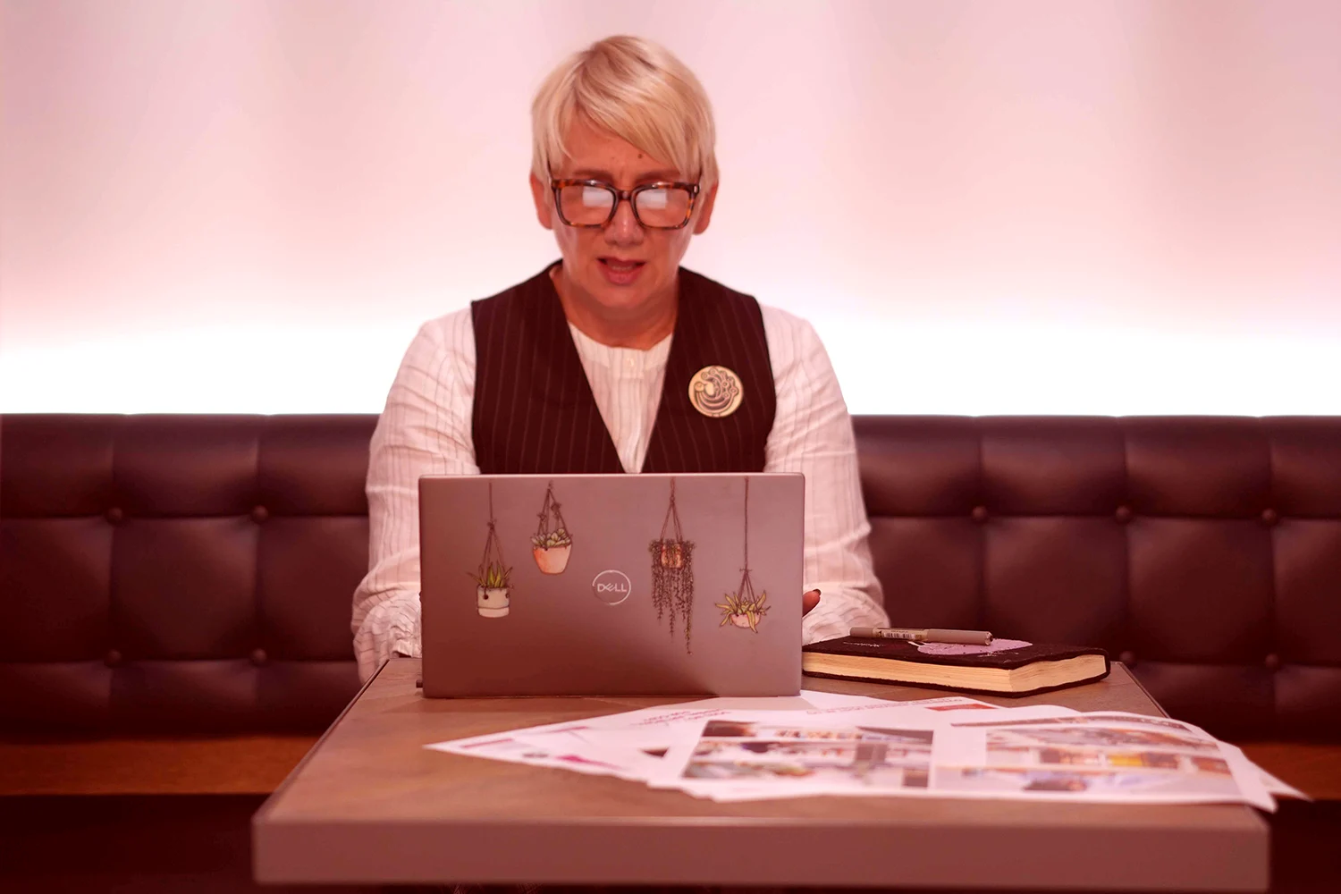 Person with short blonde hair and glasses working on a laptop with plant stickers, sitting at a table with documents and a notebook.
