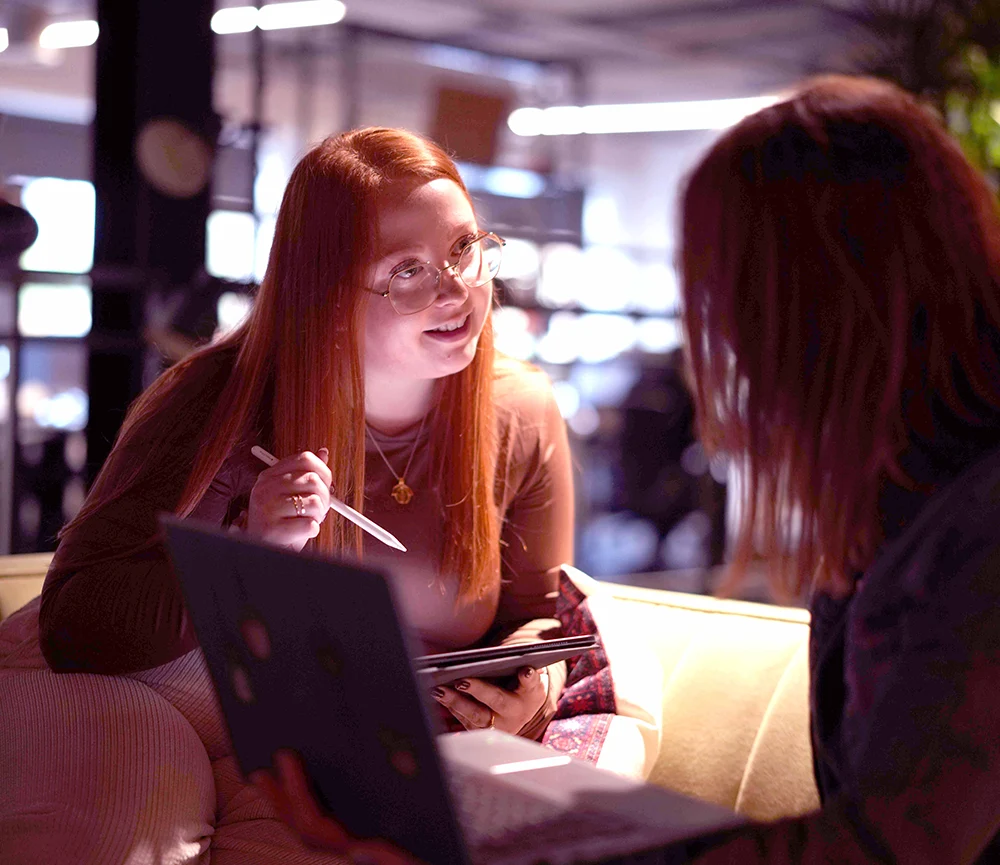Two women sitting on a couch engaged in a conversation, one holding a stylus and tablet, the other using a laptop.