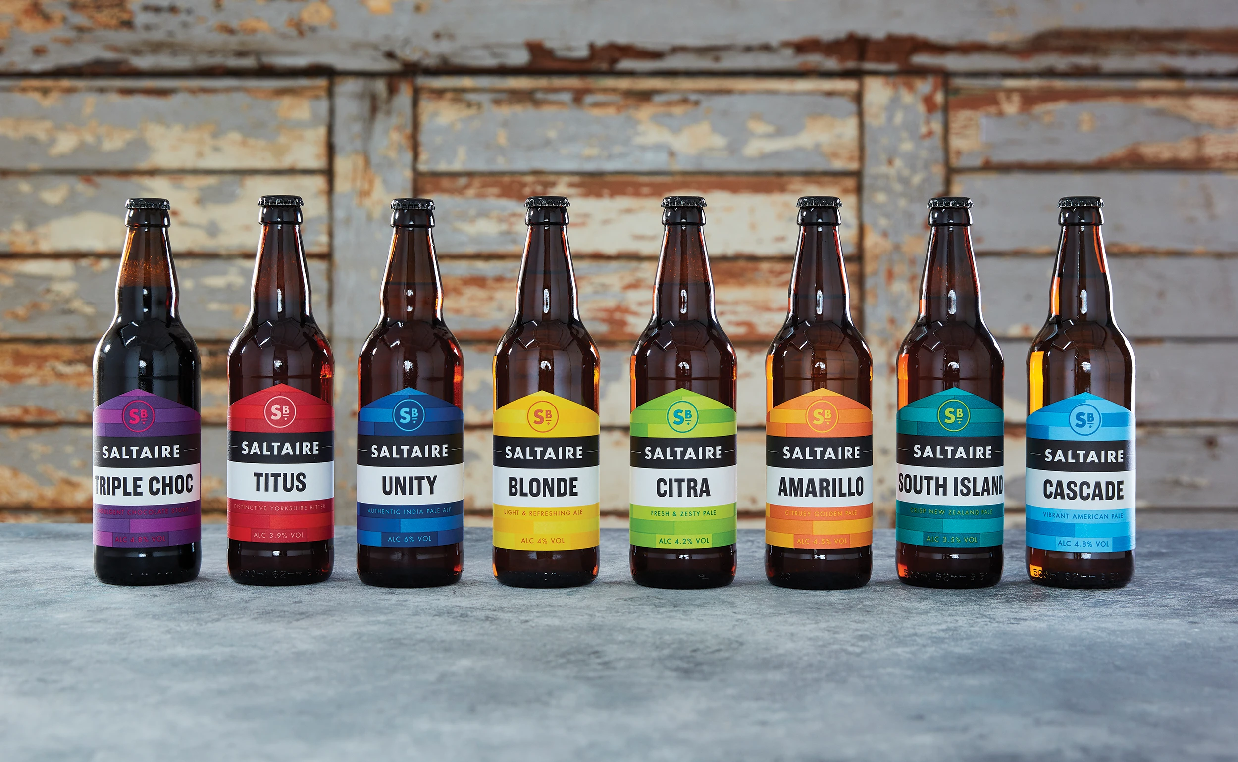 A cohesive and striking brand identity refresh for Saltaire Brewery’s core bottle range. The design features a bold, geometric "shield" label system with vibrant colour-coding for each variant—including Triple Choc, Titus, and Cascade—to ensure clear range navigation and maximum shelf standout. The minimalist "SB" monogram and clean, sans-serif typography modernise the heritage brand while maintaining its craft-led roots, all presented against a rustic, weathered wooden backdrop.