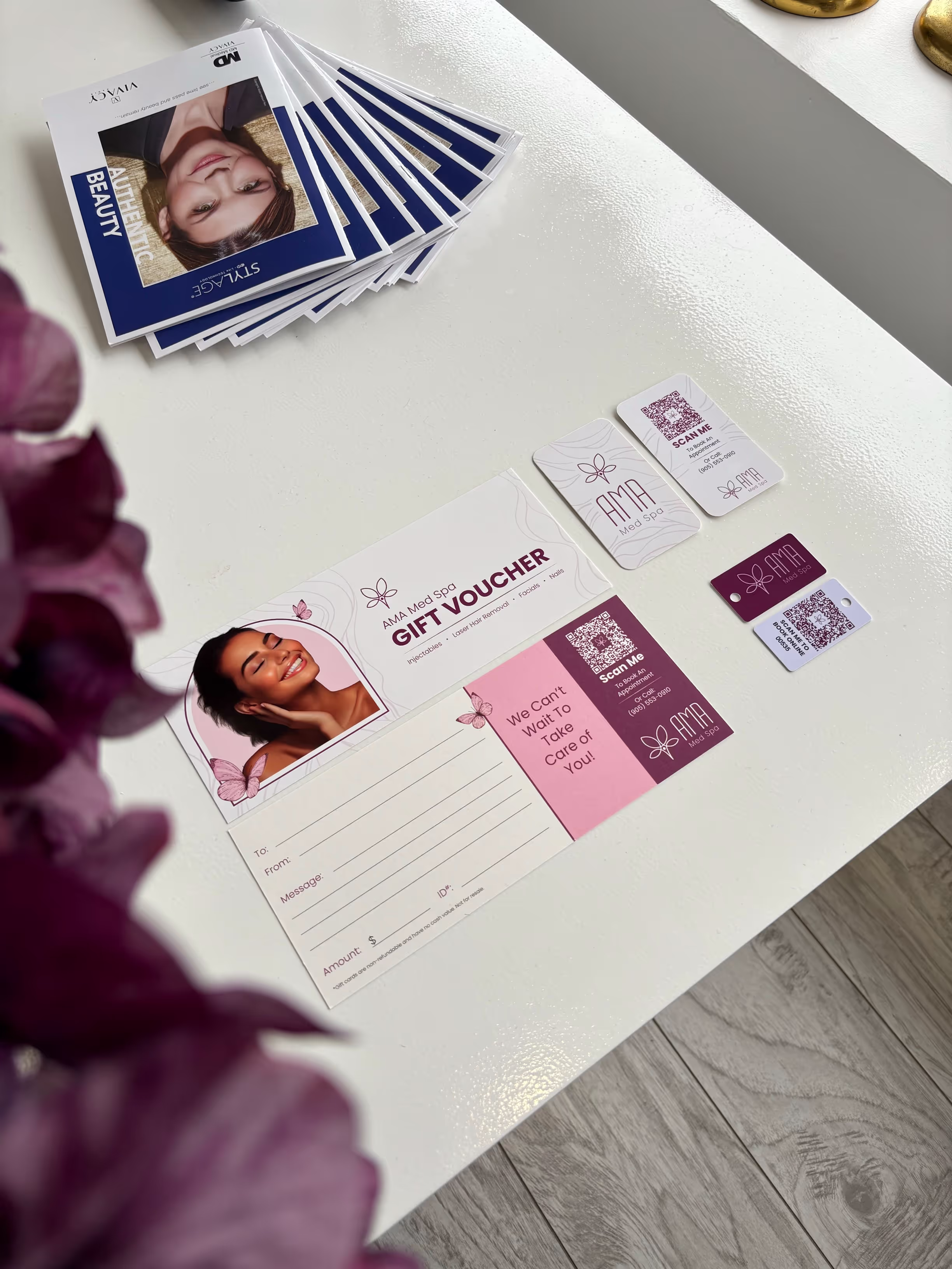 A variety of marketing materials for a spa or wellness business called AMA Med Spa. In the center is a gift voucher with a smiling woman's face on it. Around the gift voucher are various accessories like QR code business cards and stickers with the AMA logo, which appears to be a butterfly motif. On the left side, there are multiple copies of what looks like a product or service brochure featuring a woman's closeup portrait on the cover. The overall aesthetic is sophisticated and feminine, using shades of pink, purple, and blue. Several small purple flowers are scattered around the table, adding a natural, organic touch to the scene.
