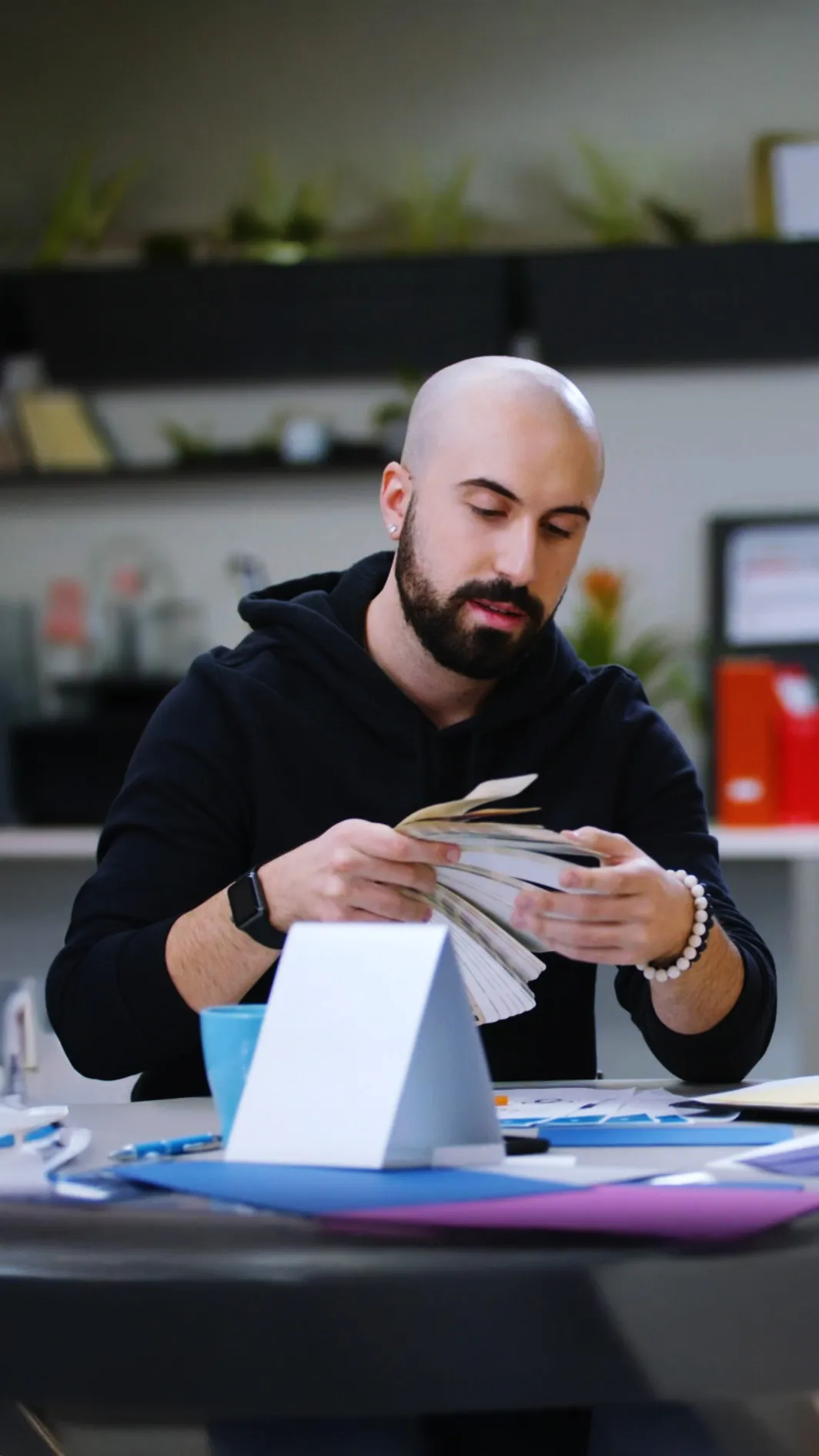 Man with a beard and black hoodie examining Pantone Colours at a desk, surrounded by office supplies and folders.