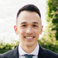 Smiling man with short dark hair wearing a suit and tie outdoors with greenery in the background.