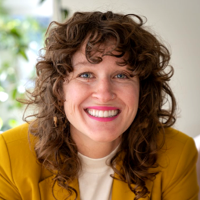 Smiling woman with curly brown hair wearing a yellow blazer and white top in a bright room.
