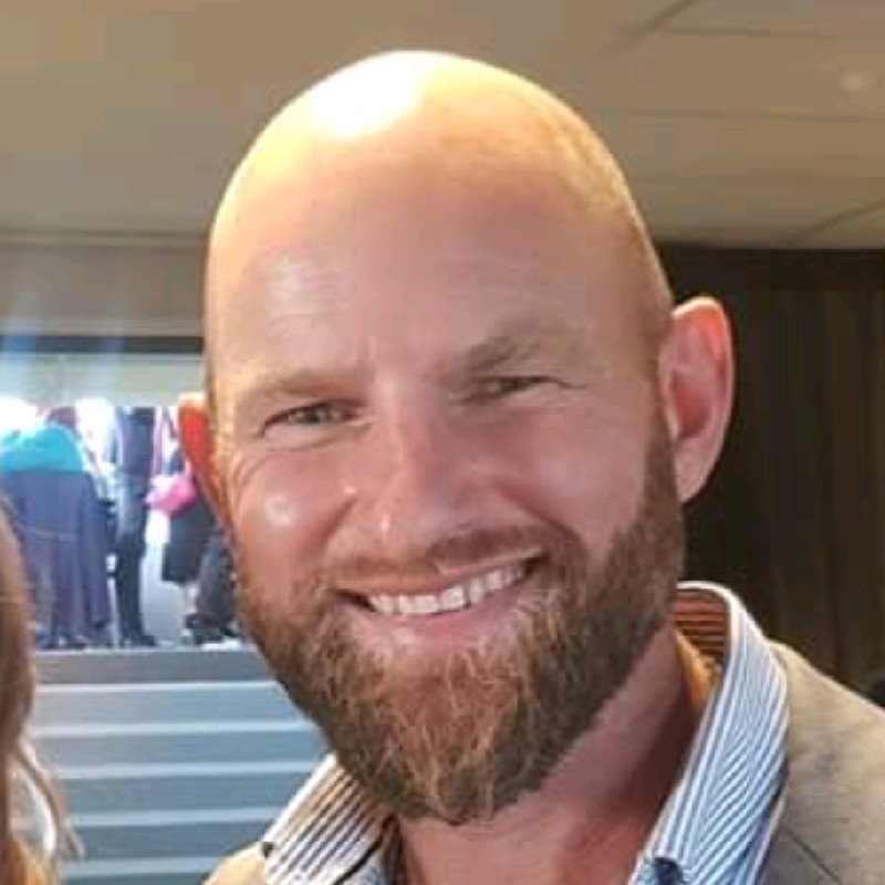 Smiling bald man with a beard wearing a striped shirt and jacket indoors.