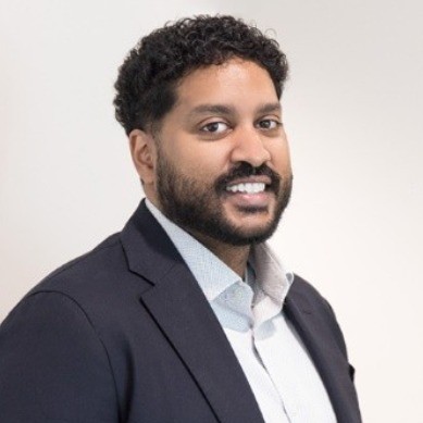 Smiling man with curly hair and beard wearing a dark blazer and light collared shirt against a plain light background.