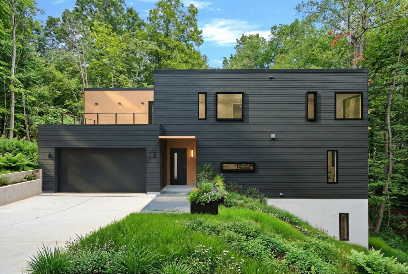 Modern two-story house with black horizontal siding, multiple rectangular windows, a large black garage door, and lush green landscaping in front.