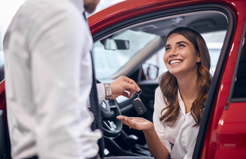 A woman seated in a red car smiles as she receives the keys from the dealership's leasing manager.