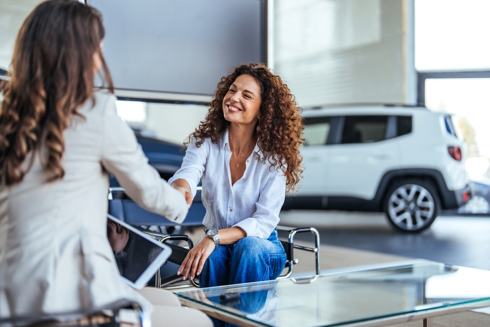 A smiling woman with curly hair shakes hands with the leasing manager at a car dealership.