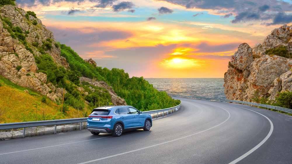 A blue SUV drives along a coastline highway at sunset