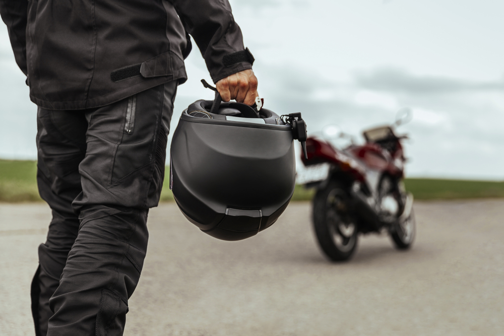 A man in a leather jacket walks toward his parked motorcycle while carrying his helmet.