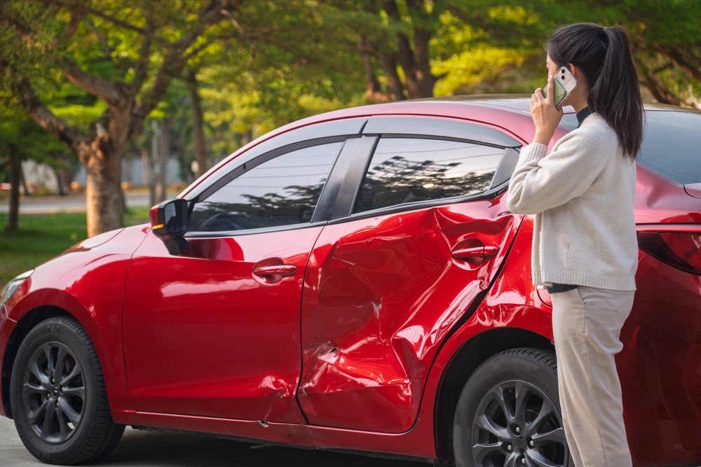 A woman on the phone stands next to a red car damaged from an accident