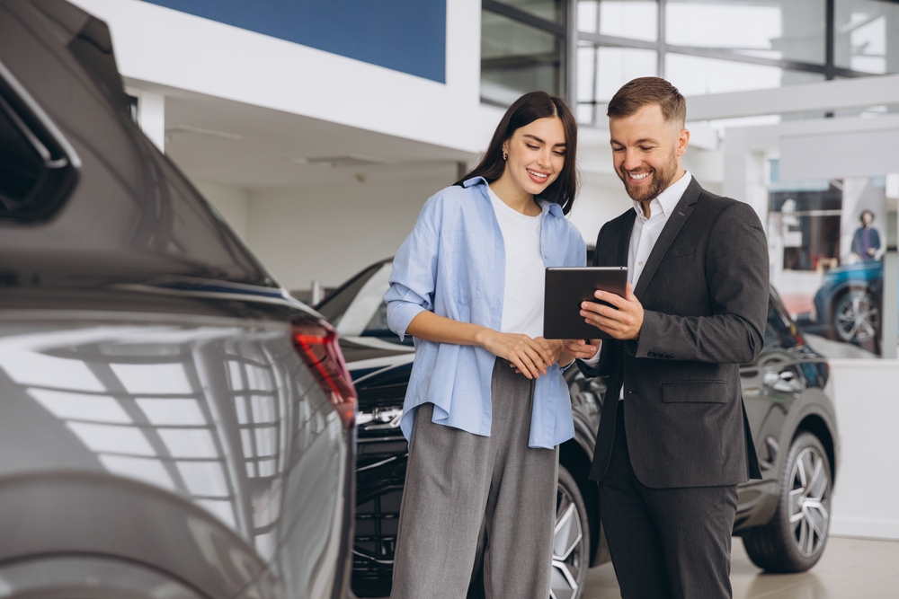 Woman stands next to car salesman inside car dealership, reviewing pre-approval paperwork on a tablet.