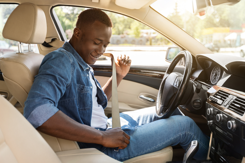 A young man in a denim shirt fastens his seatbelt in the driver’s seat of his leased car.