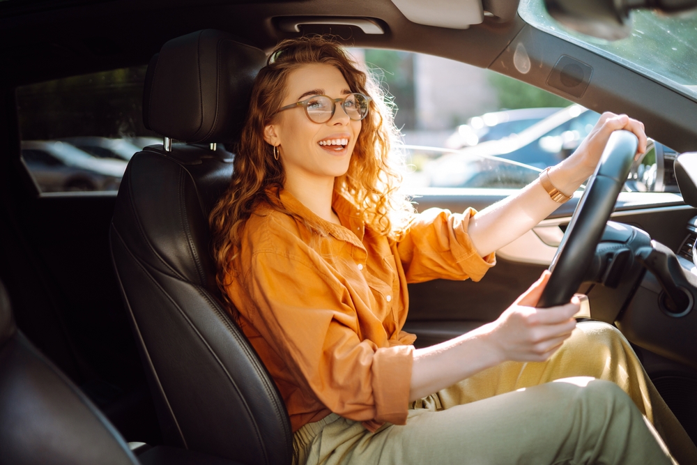 A young woman wearing glasses sits behind the wheel of her new car.