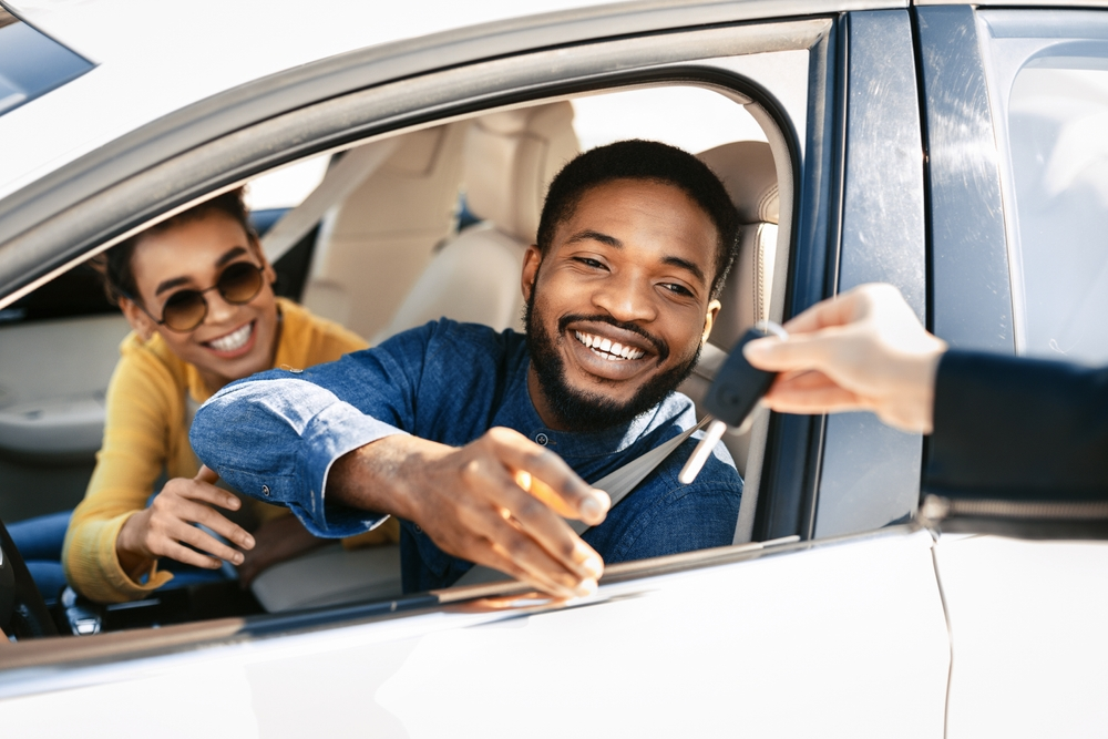 A man in a blue shirt is seated behind the while of a car, smiling with a woman seated in the passenger seat beside him.