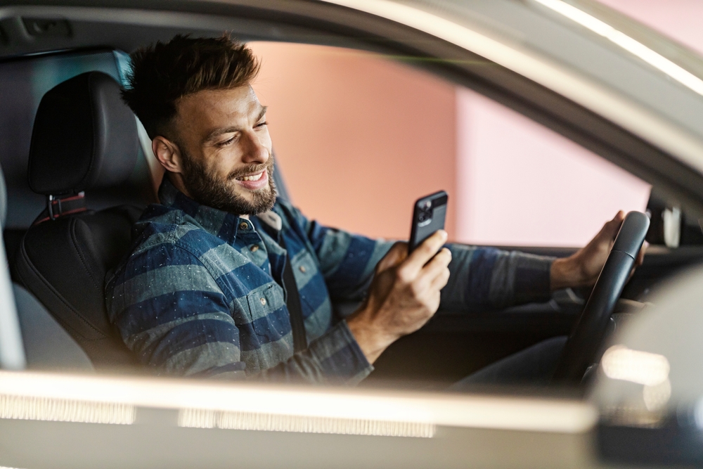 A man holds the wheel of a Tesla with one hand while looking at his phone in the other.
