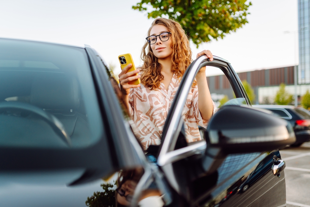  A woman with glasses stands inside the open door of a Kia while looking at her phone.