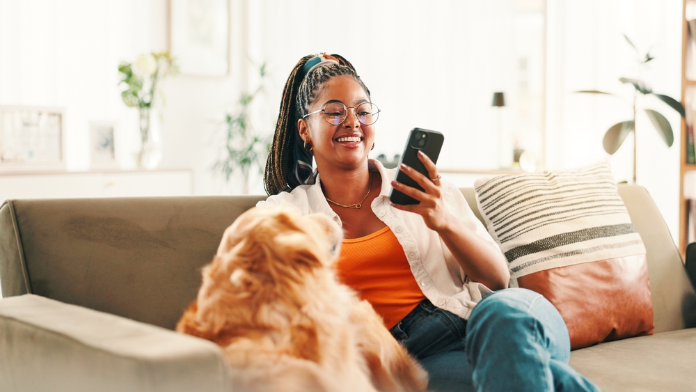 A woman wearing glasses snuggles with her dog on the couch while she checks her credit report on her phone.