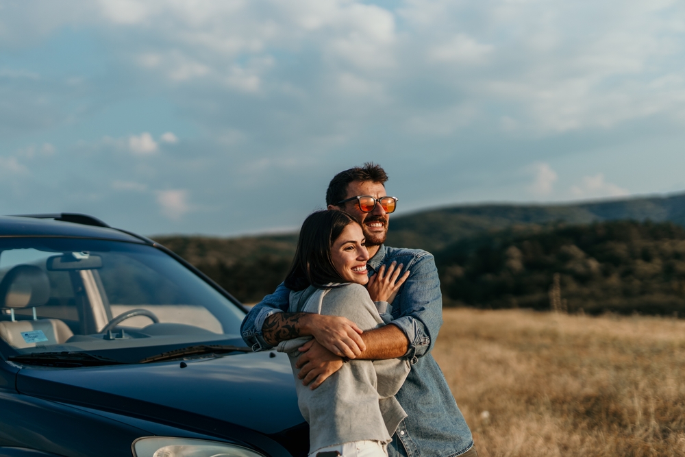 A cheerful young couple smiles as they hug in front of their car.