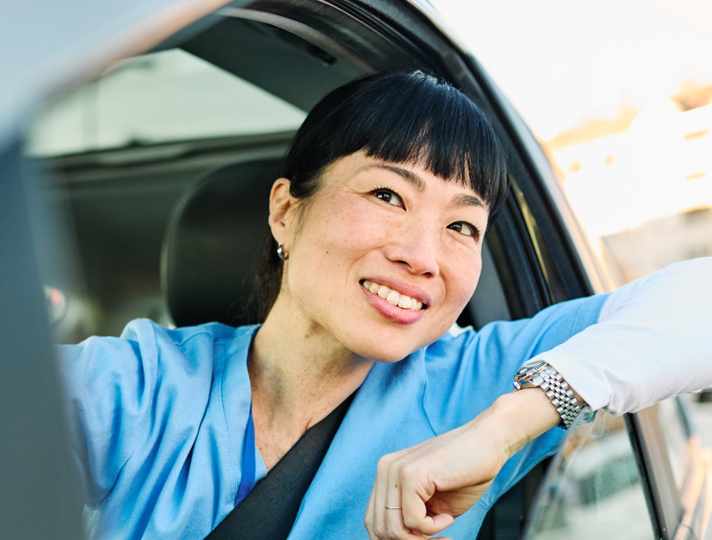 A woman with black hair smiles out the window from the driver’s seat of her car.