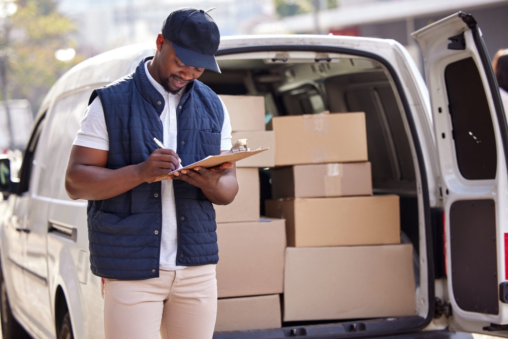 A man in a blue vest and hat holds a clipboard as he stands beside a white delivery van full of boxes.