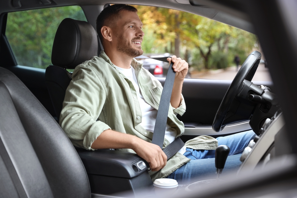 A young man in a green shirt sits in the front seat of a car, buckling his seatbelt.
